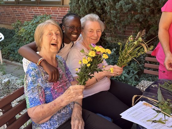 Drei Frauen, zwei ältere Damen und eine jüngere Frau, sitzen eng beieinander auf einer Bank im Freien. Alle halten kleine Blumensträuße in den Händen und strahlen vor Freude. Die jüngere Frau hat ihren Arm liebevoll um eine der Seniorinnen gelegt.