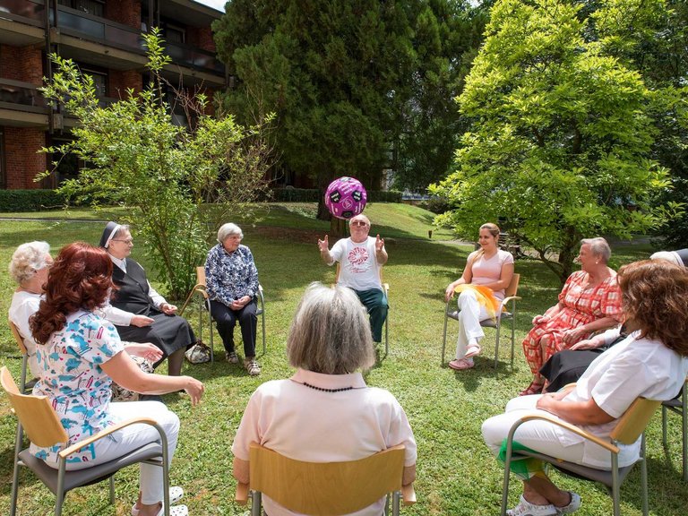 Bewohner/innen, Pflegerinnen und Ordensschwestern sitzen im Garten auf Stühlen im Kreis und werfen sich einen Ball zu