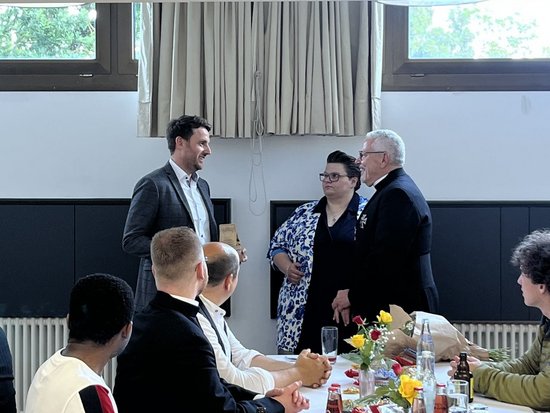 Ein geselliges Beisammensein in einem hellen Raum. Im Hintergrund unterhalten sich drei stehende Personen, während andere Gäste an einem langen Tisch mit weißen Tischdecken, Blumen und Getränken sitzen.