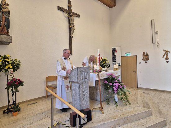 Ein Priester in weißen Gewändern steht an einem steinernen Lesepult in einer Kirche und liest vor. Im Hintergrund steht ein weiterer Mann am Altar unter einem großen Kreuz an der Wand. Blumen schmücken den Altarraum.