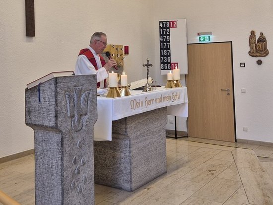 Ein Priester steht an einem Altar in einer Kirche und feiert die Messe. Hinter ihm hängt ein großes Kruzifix an der weißen Wand. Im Vordergrund befindet sich ein steinernes Lesepult, und rechts sind weitere religiöse Figuren zu sehen.