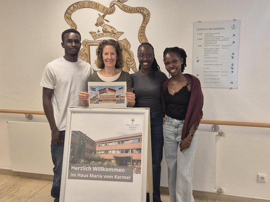 Vier Personen stehen in einem Flur vor einem Schild mit der Aufschrift „Herzlich Willkommen im Haus Maria vom Karmel“ und einem Bild des Gebäudes. Eine Frau hält eine Broschüre. Im Hintergrund ist ein Wappen an der Wand zu sehen.