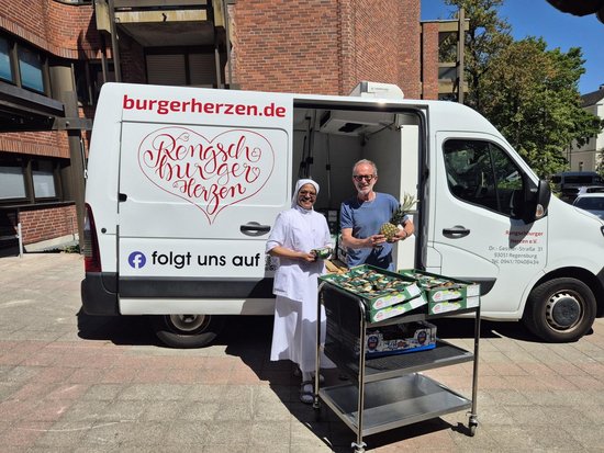 Ein weißer Lieferwagen der Organisation „burgerherzen.de“ steht im Hintergrund. Eine Nonne und ein Mann stehen davor, halten frisches Gemüse in den Händen und stehen neben einem Wagen, der mit Lebensmitteln beladen ist.